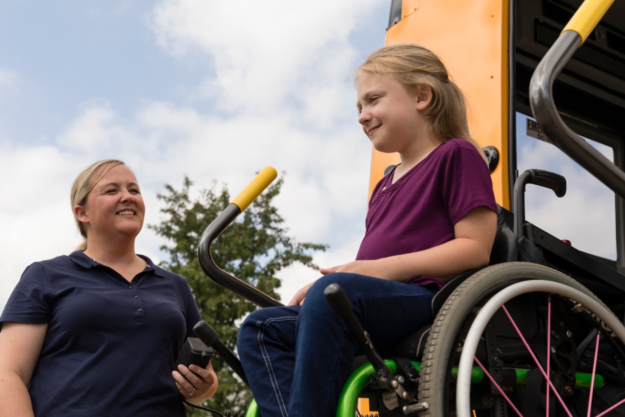 a little girl in a wheelchair exiting a school bus with the assistance of a wheelchair lift and a care attendant 