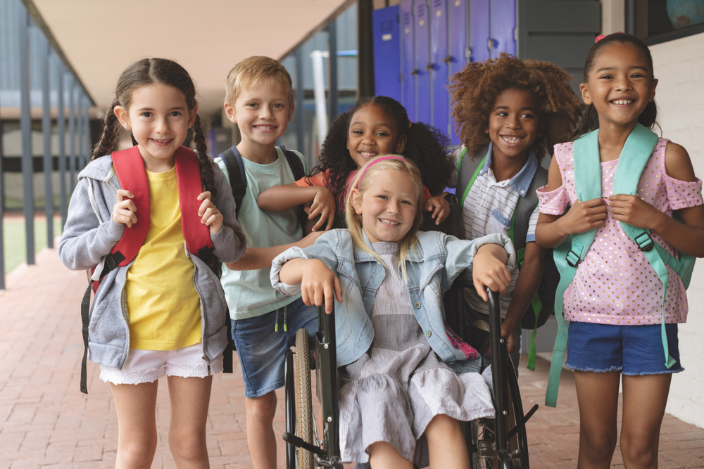 Front view of happy diverse school kids standing in outside corridor at school while a Caucasian schoolgirl is sitting on wheelchair in foreground