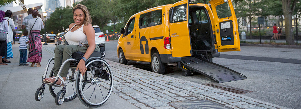 woman in wheelchair on sidewalk outside of a wheelchair accessible taxi
