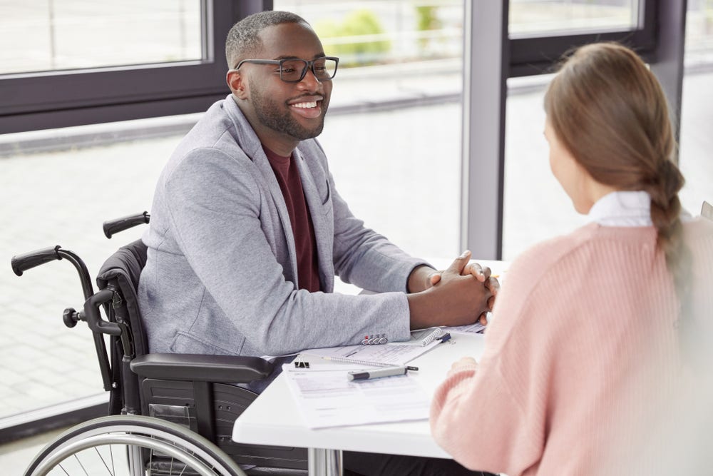 Handsome cheerful black male in wheelchair involved in charity, meets with female investor, discuss main details, helps physically challenged people. People, health and business concept Handsome cheerful black male in wheelchair involved in charity, meets with female investor, discuss main details, helps physically challenged people. People, health and business concept