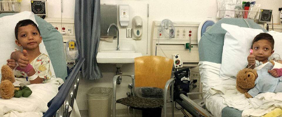 Two young brothers sit in adjacent hospital beds and give a thumbs up