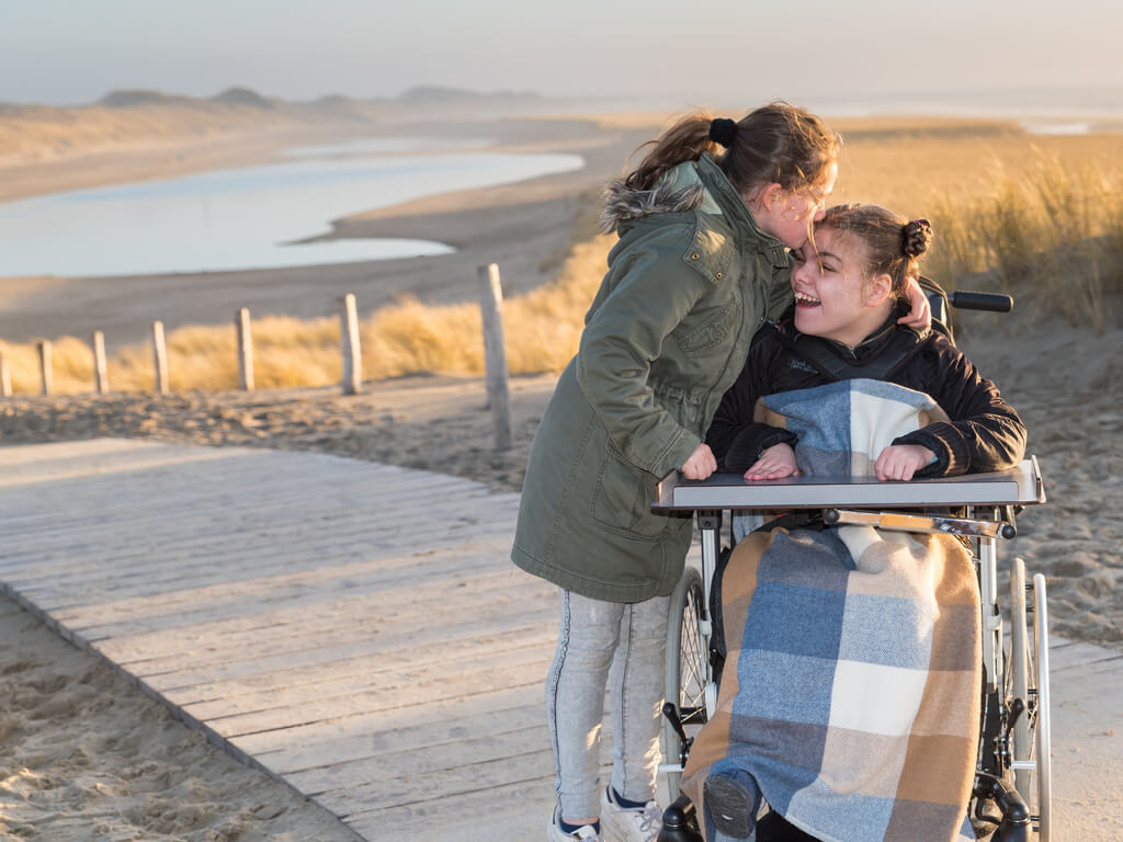 A young girl kisses the head of her sister who is in a wheelchair on a boardwalk