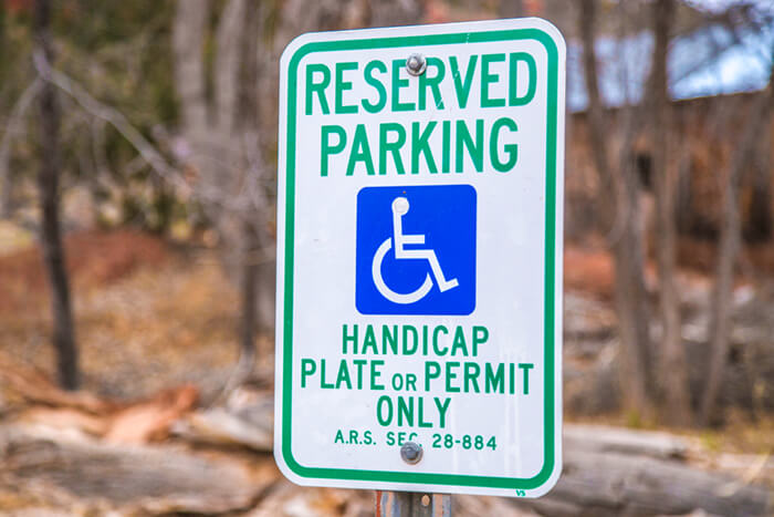 A sign for handicap parking indicates the need to have a handicap parking permit to avoid a fine|A man in a wheelchair sits next to a vehicle with the driver's door ajar illustrating a handicap parking permit requirement|A blue handicap parking permit hangs from a rear-view mirror