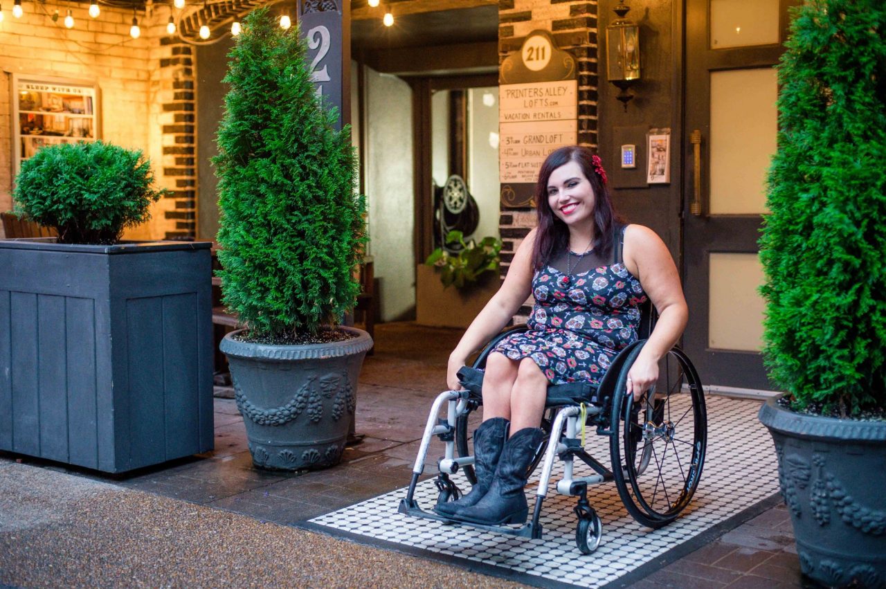 Deanna is sitting outside of a hotel in a wheelchair. Deanna is wearing a pretty dress with boots.