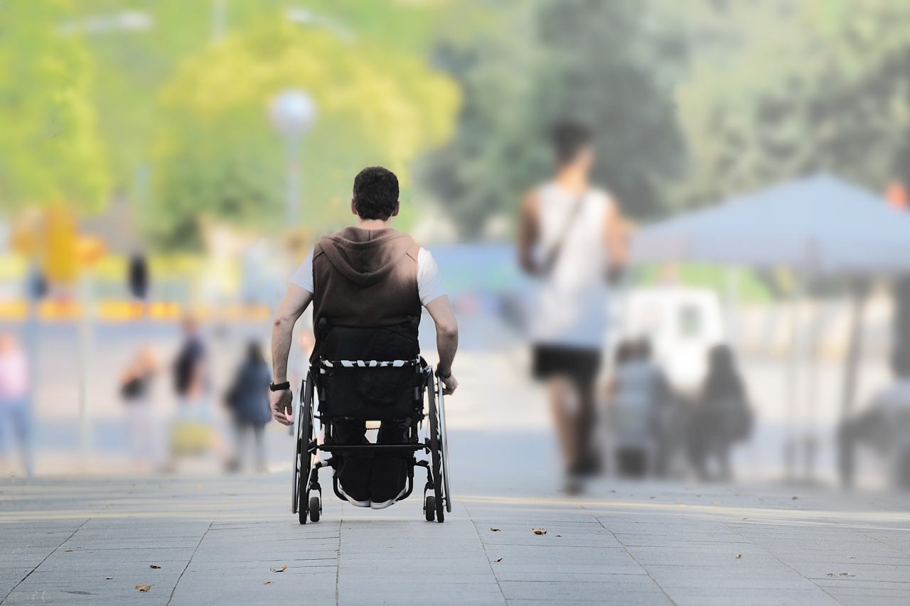 ALS affected young man on a wheel chair in the street among other people.