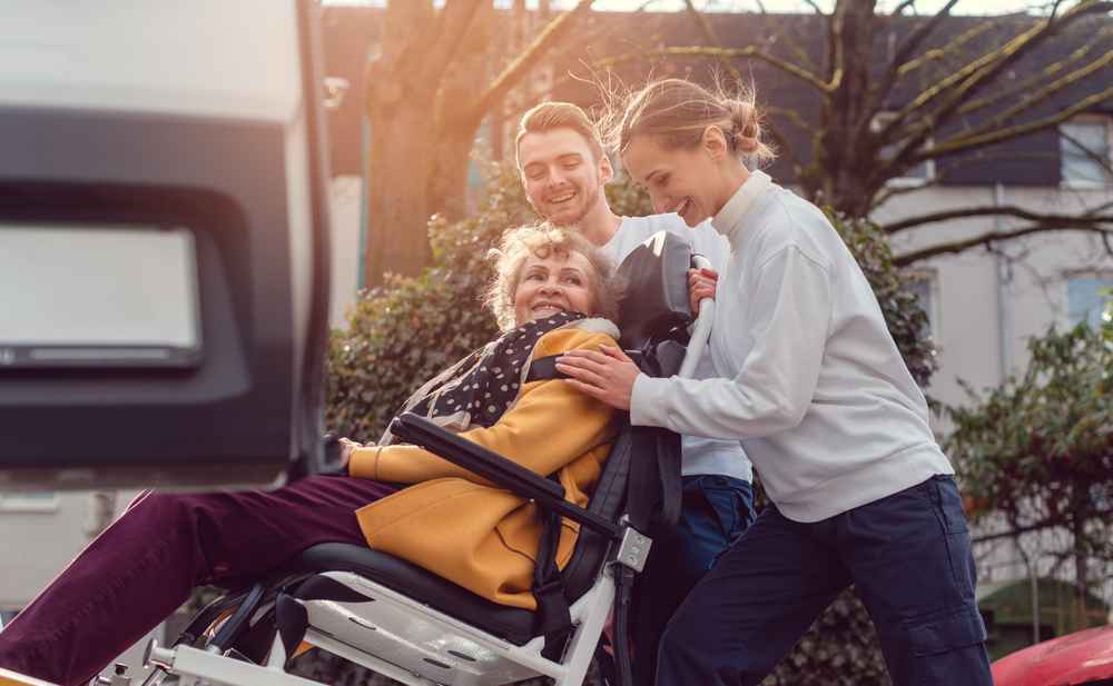 two individuals who work for a wheelchair van service helping another person up a wheelchair ramp