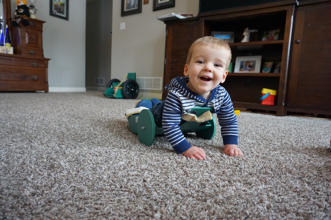 Brody, a toddler, smiles at the camera while using The Frog device on soft carpet