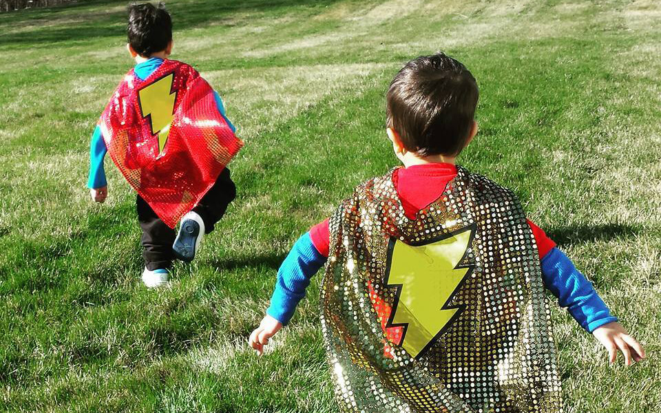 Two young boys run away from the camera with superhero capes on in a field. Two young boys run away from the camera with superhero capes on in a field.