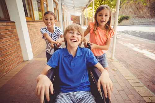 a young boy wearing a blue shirt in a wheelchair is being pushed by 2 girls who are is friend